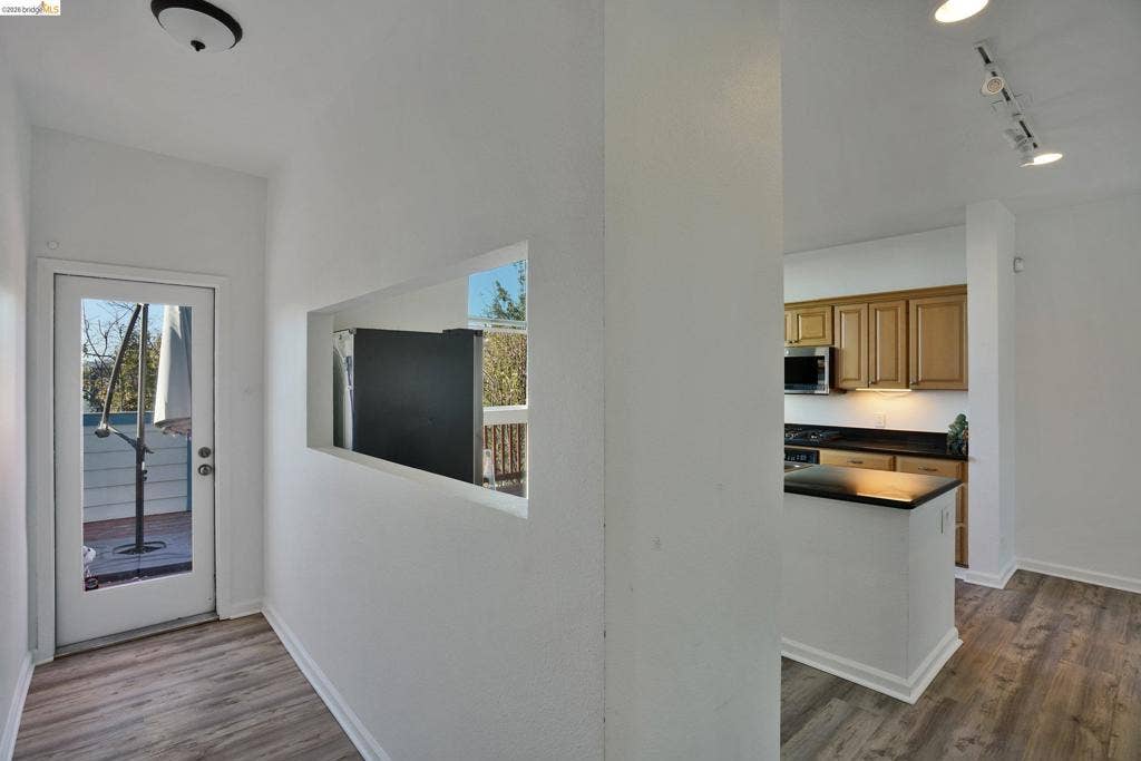 Interior featuring wood-finish flooring, recessed pass-through window, full-light entry door, wooden cabinetry, and dark solid-surface countertops