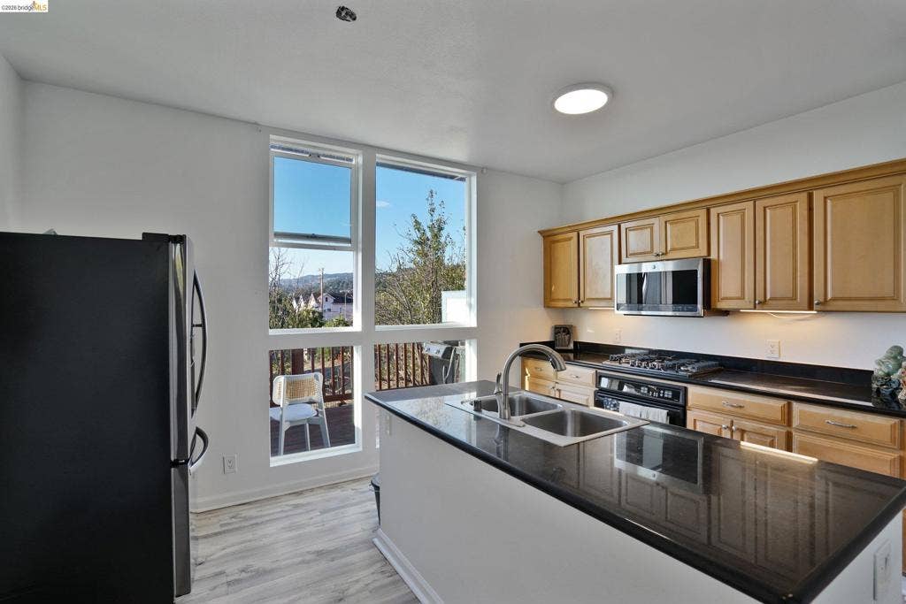 Kitchen featuring wood cabinetry, dark stone-finish countertops, stainless steel appliances, a central island with a double-basin sink, and floor-to-ceiling windows