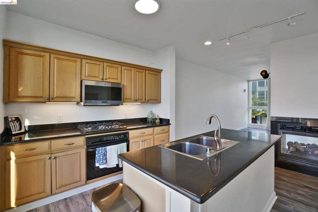 Kitchen featuring natural wood cabinetry, dark stone countertops, stainless steel gas cooktop with built-in microwave, center island with double-basin stainless steel sink, and wood-finish flooring