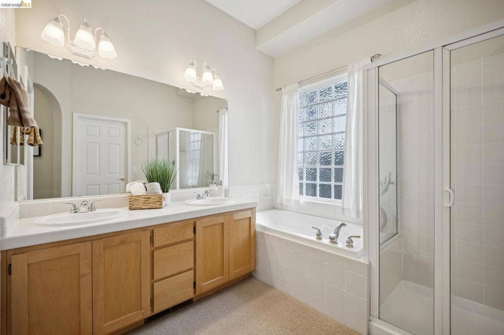 5503 Rainbow Cir, Santa Rosa, CA 95409 Dual-sink vanity with light-tone wood cabinetry, porcelain-tiled tub deck, glass-enclosed shower, and textured glass window