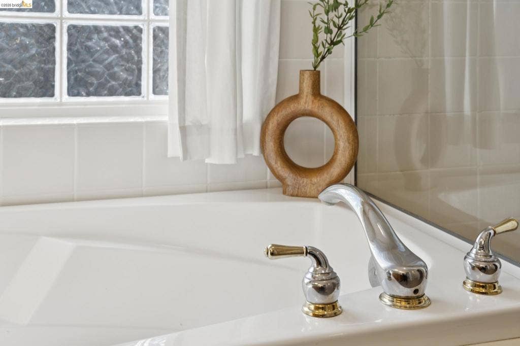 5503 Rainbow Cir, Santa Rosa, CA 95409 Soaking tub featuring a white acrylic finish, deck-mounted chrome faucet with gold-tone accents, square tile surround, and an integrated glass block window