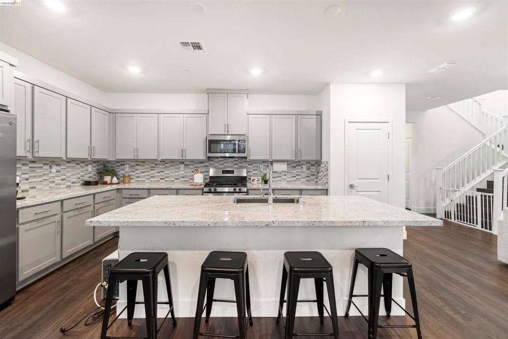 539 Gemstone Way, Oakley, CA 94561 Kitchen featuring light grey cabinetry, an expansive center island with a stone countertop, a multi-toned tile backsplash, stainless steel appliances, and dark wood-finish flooring