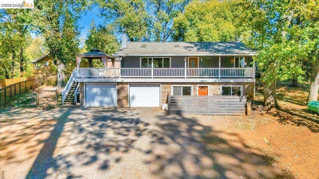 5827 Lookout Ct, CA 95233 Split-level exterior featuring a covered deck with white railing, double-bay garage, horizontal privacy fencing, and mature surrounding trees