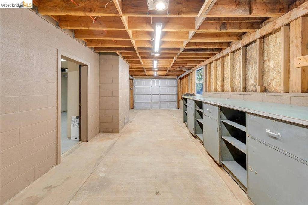 5827 Lookout Ct, CA 95233 Garage space featuring exposed ceiling joists, concrete block walls, overhead linear lighting, a roll-up garage door, and built-in metal cabinetry