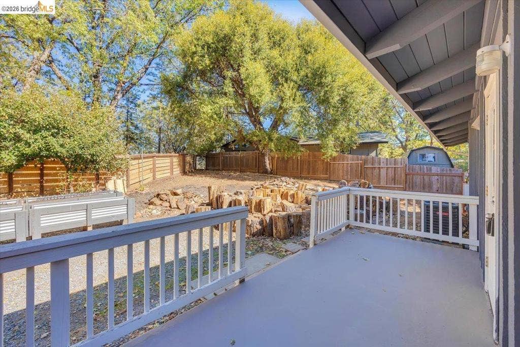 5827 Lookout Ct, CA 95233 Covered porch featuring a painted wood floor and white railing, opening to a fenced backyard with horizontal wood privacy fencing, raised metal garden beds, and mature trees
