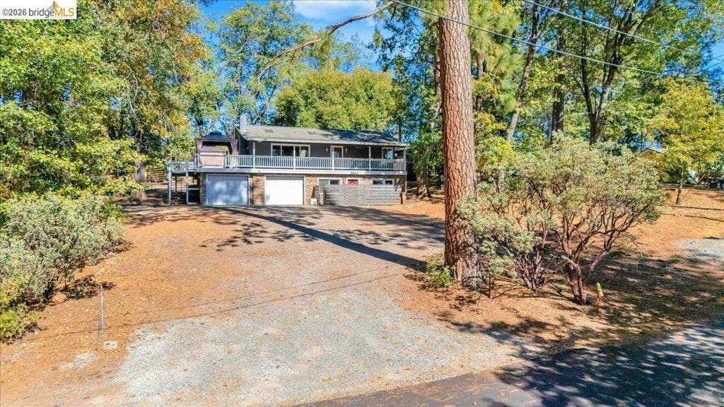5827 Lookout Ct, CA 95233 Multi-level exterior featuring a wide upper-level deck, two-car garage, gravel driveway, and mature trees on a wooded lot