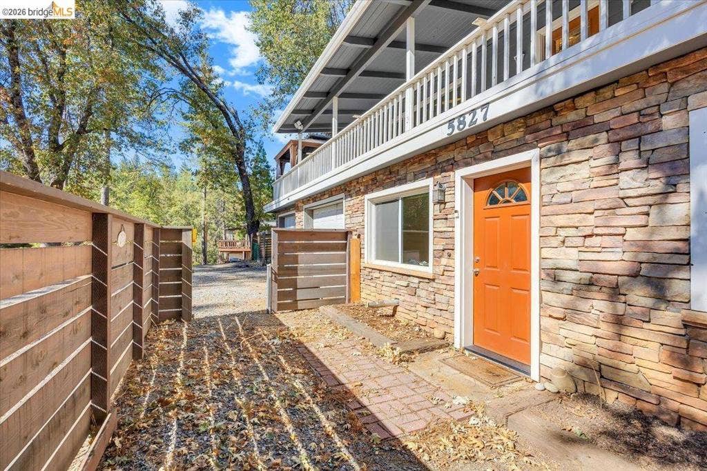 5827 Lookout Ct, CA 95233 Exterior facade featuring multi-tonal stacked stone siding, a covered upper-level balcony with metal railing, and horizontal wood privacy fencing