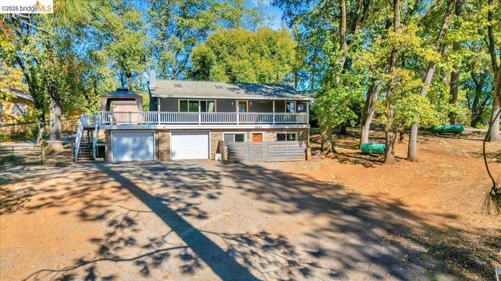 5827 Lookout Ct, CA 95233 Split-level exterior featuring a full-width upper deck, two-car garage, brick foundation detailing, and a slatted privacy screen on a lot with mature trees