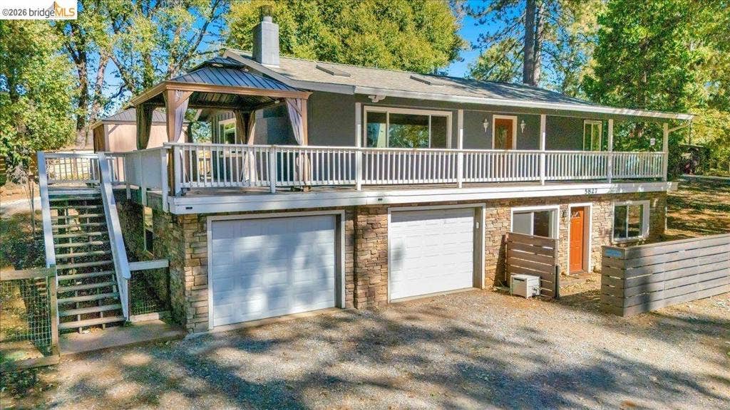 5827 Lookout Ct, CA 95233 Split-level exterior featuring a stone veneer lower level, elevated wrap-around deck with white railing, double-garage configuration, and staircase access