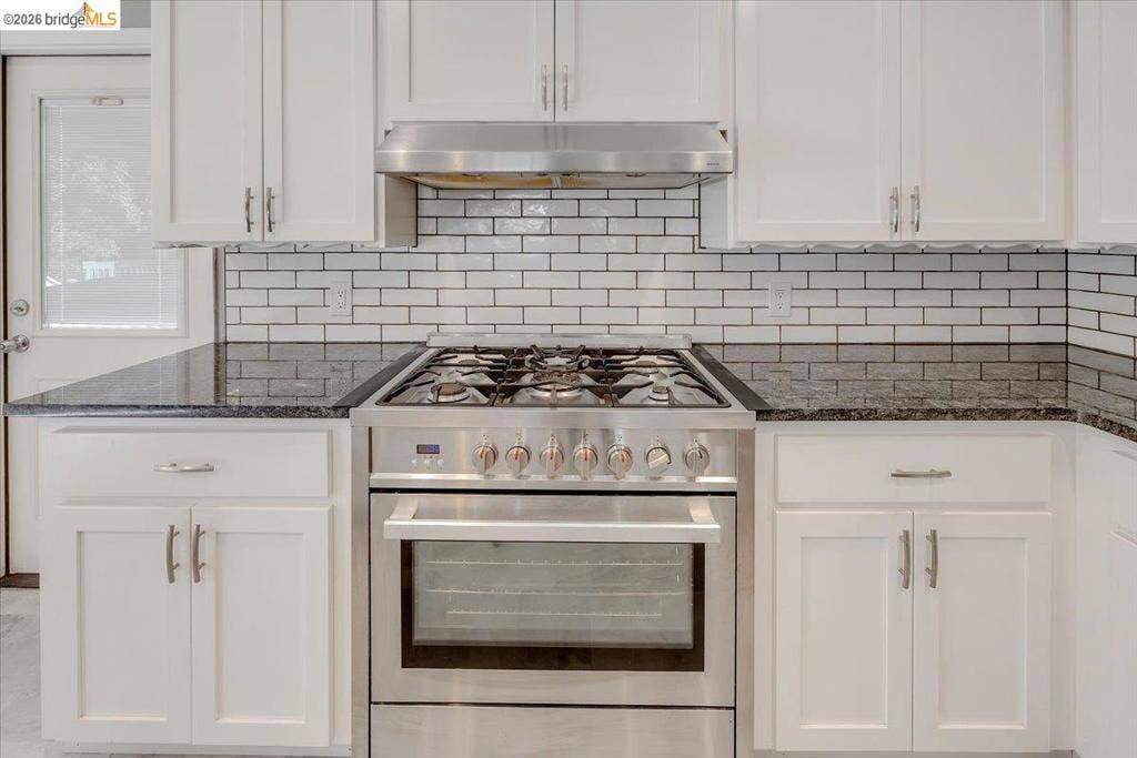 5827 Lookout Ct, CA 95233 Kitchen featuring white Shaker-style cabinetry, dark stone countertops, a subway tile backsplash, and a stainless steel gas range with a matching overhead hood