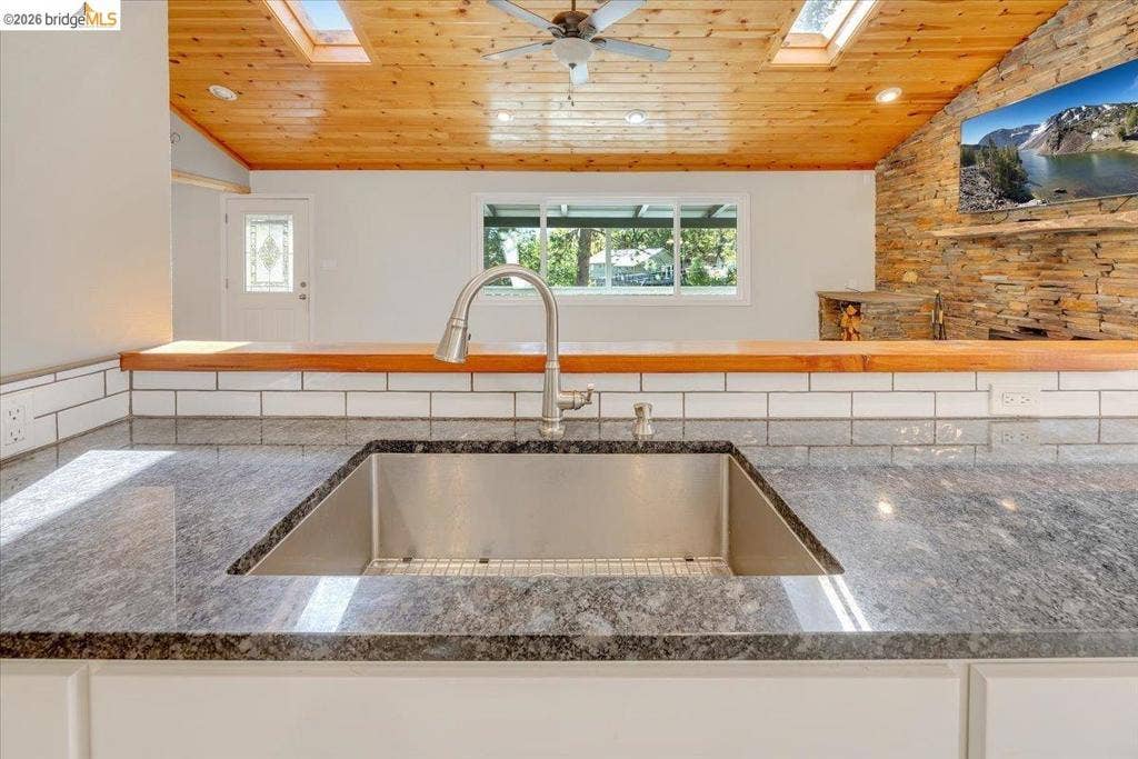 5827 Lookout Ct, CA 95233 Kitchen island featuring a stone countertop, under-mount stainless steel sink, and high-arc faucet, set against a white subway tile backsplash, wood-plank ceiling, and stone-accent wall