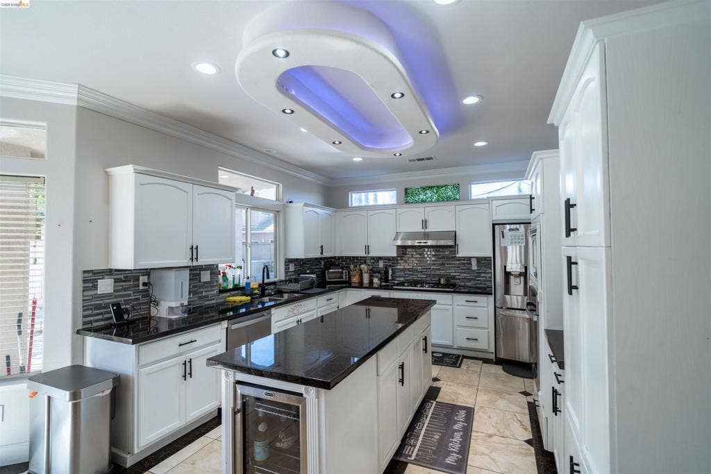 4600 Via Terreno, Modesto, CA 95357 Kitchen featuring custom white cabinetry, a center island with built-in wine refrigeration, dark stone countertops, a tiled backsplash, and an elliptical recessed ceiling feature with integrated lighting