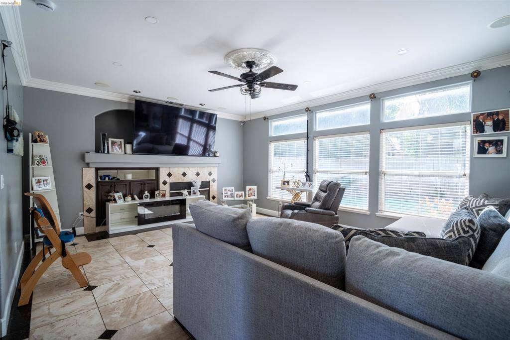 4600 Via Terreno, Modesto, CA 95357 Living area featuring oversized floor tiles with decorative inlay, a fireplace with integrated cabinetry, crown molding, a central ceiling fan with medallion detail, and a wall of multi-pane windows