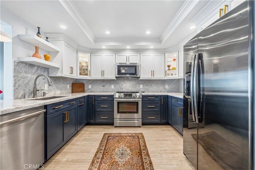Remodeled Kitchen With The beautifully upgraded kitchen is a showstopper, featuring sophisticated two-tone cabinetry with rich navy blue lowers and crisp white uppers, elegant brushed gold hardware, gorgeous blue calcite countertops with a matching full-height backsplash, and a complete suite of stainless steel appliances. Illuminated glass-front display cabinets, decorative floating shelves, recessed lighting, and an elegant tray ceiling with crown molding add architectural charm, while abundant cabinet...
