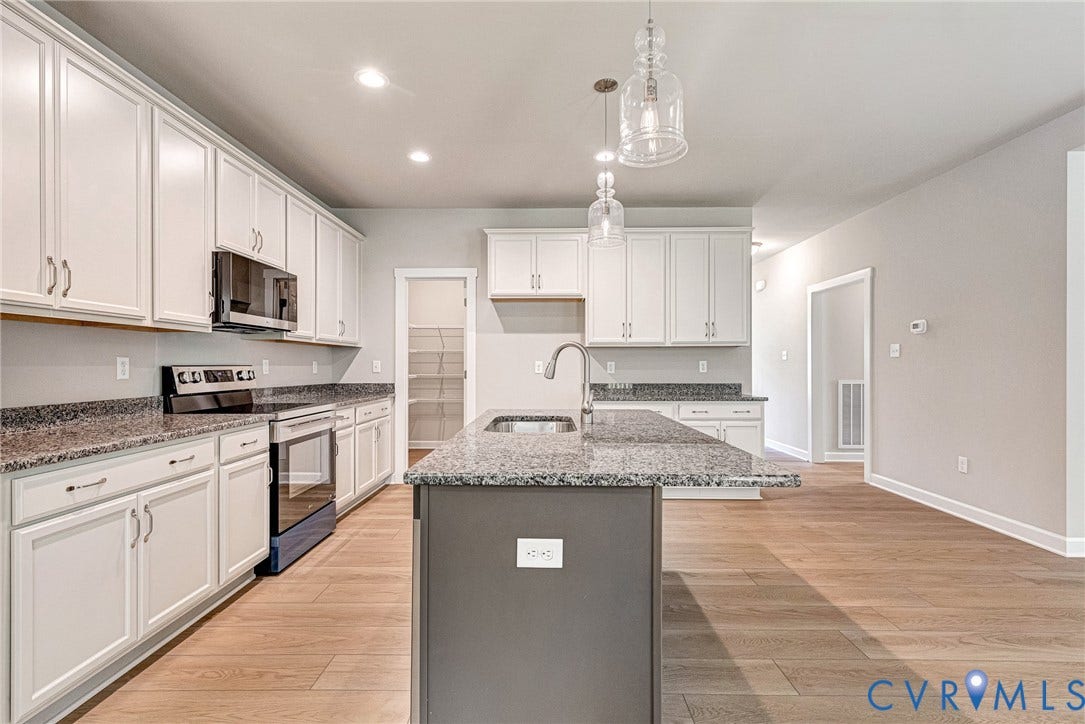 Open-concept kitchen and living area featuring white cabinetry with stainless steel appliances, granite countertops, a centered island with prep space, wood-finish flooring throughout, and pendant lighting suspended above the counter workspace
