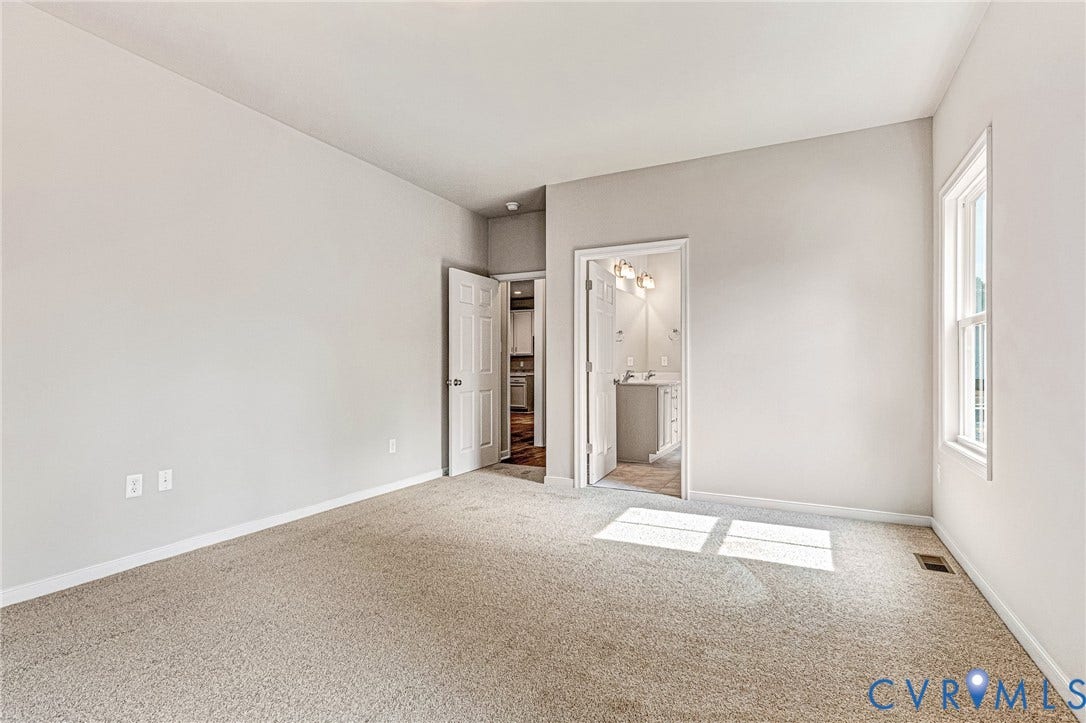 Spacious carpeted room featuring a double-hung window, white baseboards, and a doorway leading to a bathroom with a white vanity and wall-mounted light fixture