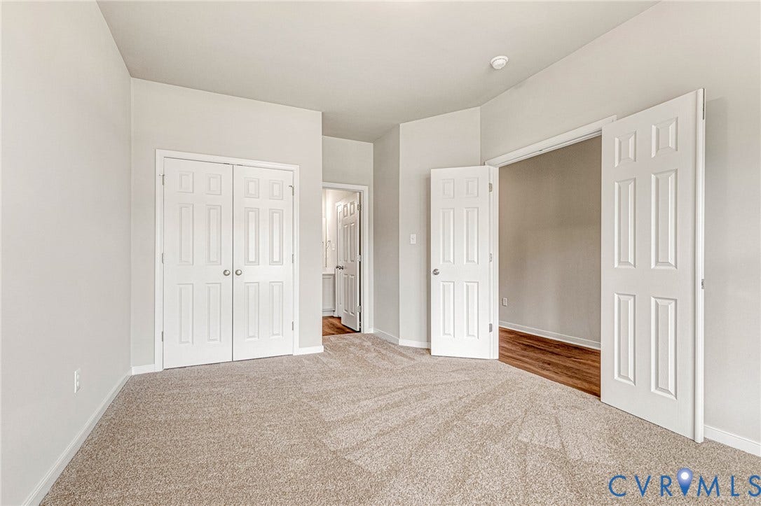 Neutral-toned room featuring carpet flooring, two paneled closet doors, and a wide entryway with double doors opening to an additional space with wood-finish flooring
