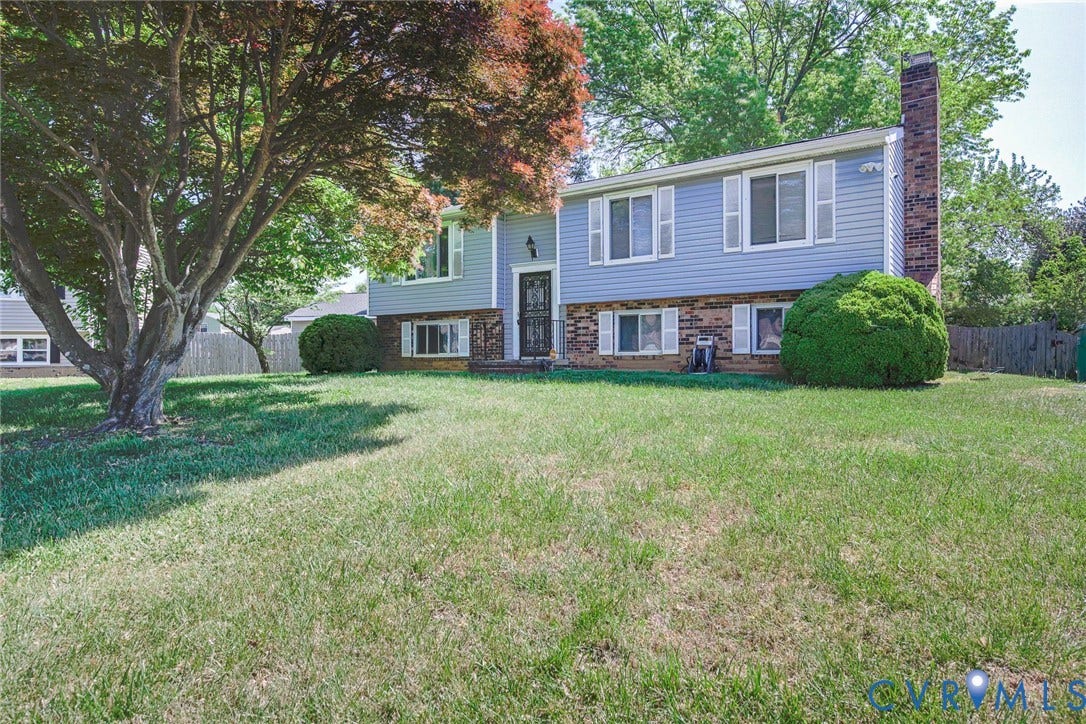 The property features a split-level design with light blue siding and a brick facade, complemented by a large front yard with a mature tree and well-maintained shrubbery