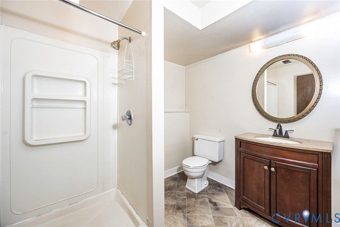 Bathroom featuring a built-in shower stall with integrated shelving, dark wood-finish vanity with a light-toned countertop, and a round mirror with an ornate frame