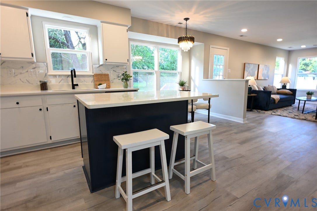 912 Harmony Road, Fairfield, VA 23059 Open-concept kitchen and living area featuring a two-tone island with waterfall countertop, white cabinetry with hardware accents, stainless steel sink, wood-finish flooring throughout, and abundant natural light from multiple windows and glass doors