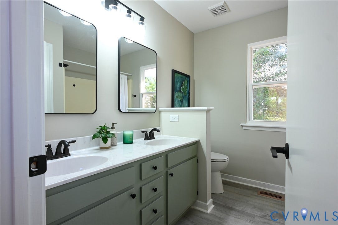 912 Harmony Road, Fairfield, VA 23059 Dual vanity bathroom with white quartz countertop, matte black hardware, framed mirrors with integrated lighting, sage green cabinetry with multiple drawers, wood-finish flooring, and abundant natural light from large windows