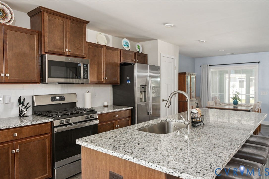 7236 George Wythe Lane, Summer Maple, New Kent, VA 23124 Kitchen featuring an expansive island with a stainless steel sink and a high-arc faucet, granite-style countertops, stainless steel appliances, wood cabinetry, and a white subway tile backsplash