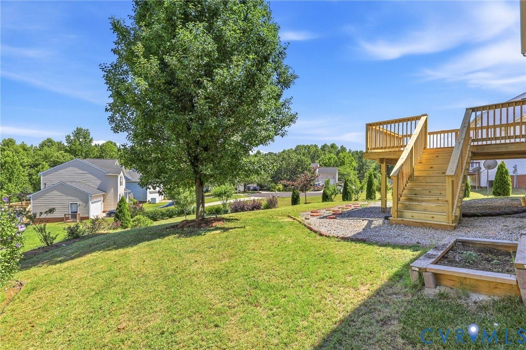 Expansive green lawn with a mature shade tree, featuring a multi-level wood deck with stairs, a gravel patio area with stepping stones, and a raised garden bed