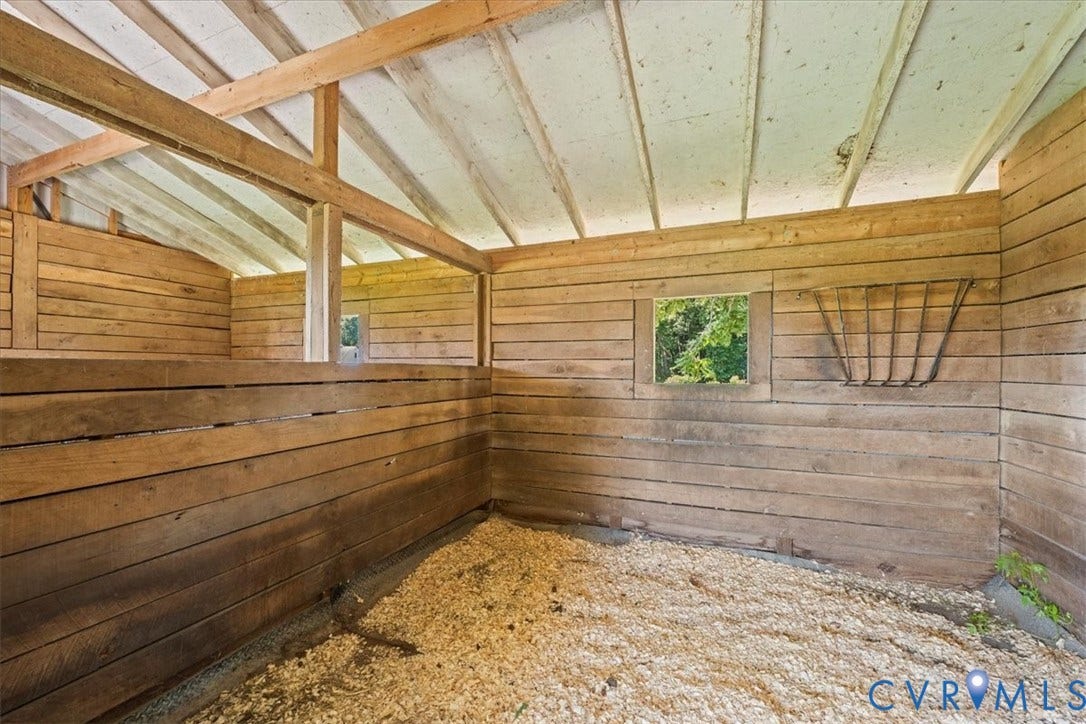 Interior stable structure featuring wood plank walls, exposed wood ceiling beams, a small window opening, and a gravel-covered floor