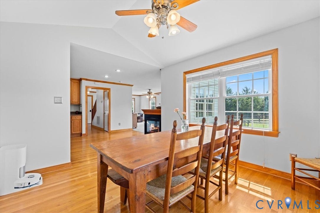 Open-concept interior featuring light wood-finish flooring, a large window with natural wood trim, vaulted ceilings, and a ceiling fan with integrated lighting