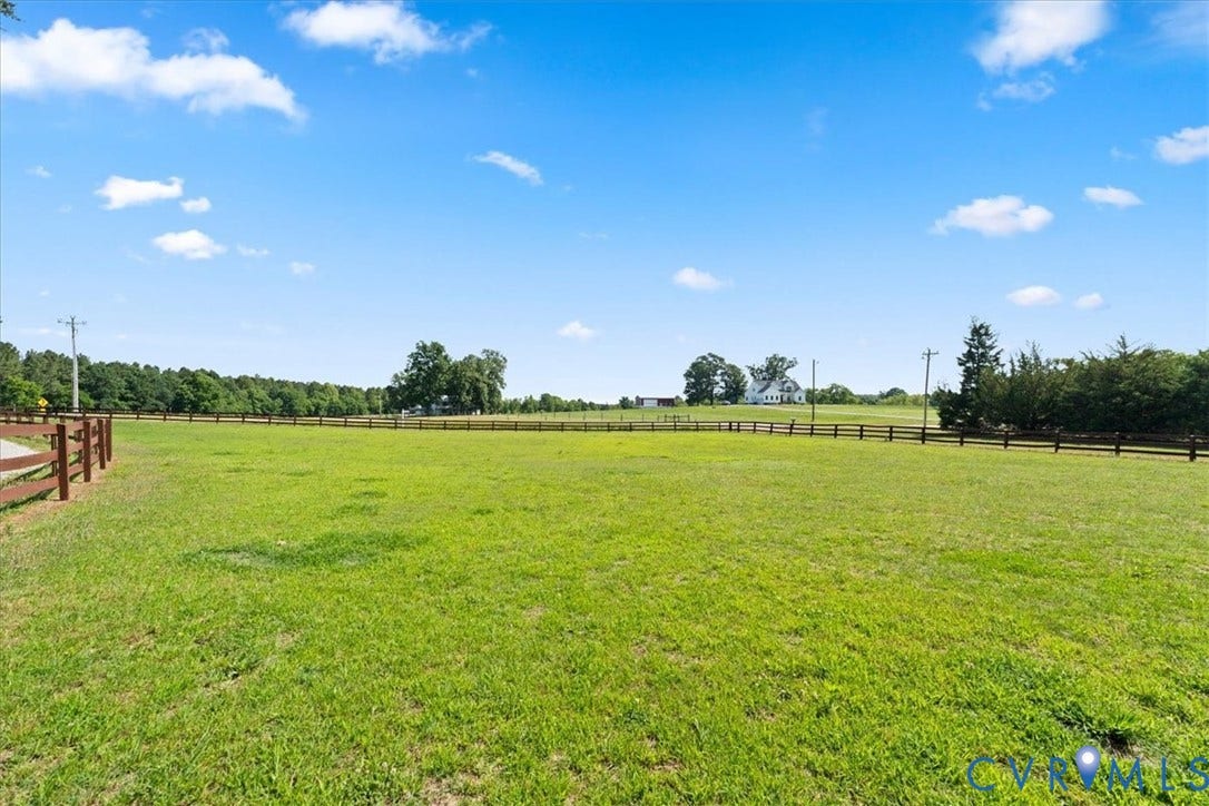 Expansive green field featuring a natural wood-toned fence line, distant trees, and a clear sky with scattered clouds
