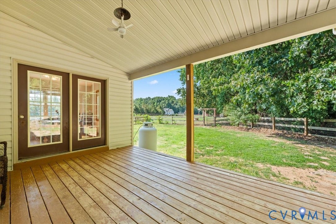 Covered wooden deck featuring a ceiling fan, dual French doors with grid-patterned glass, white vinyl siding, and a spacious green lawn bordered by a wooden fence