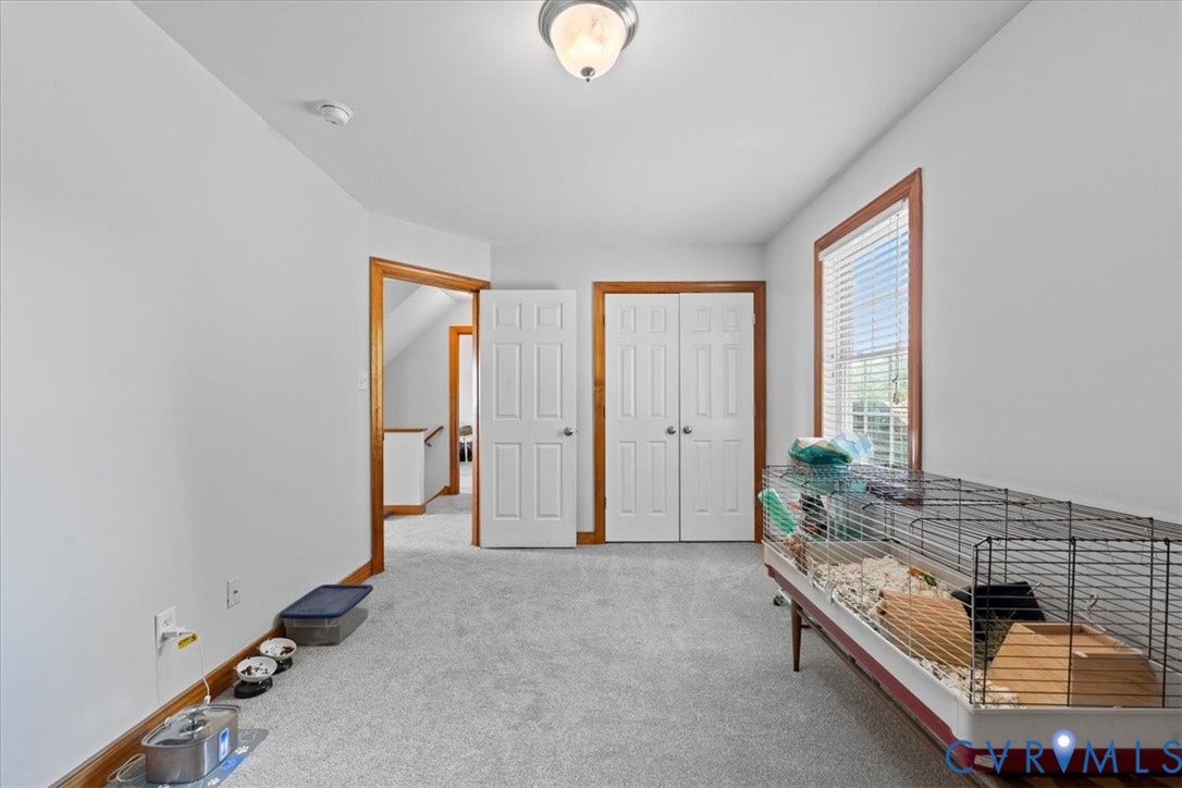 Spacious room featuring light gray carpeting, neutral wall finishes, and a single window with wooden trim