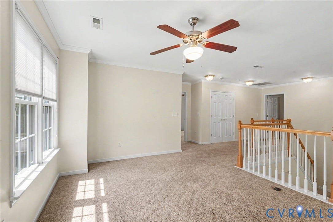 Spacious carpeted room featuring a ceiling fan with light fixture, multiple windows with blinds, white trim, and a staircase with wood and white balusters