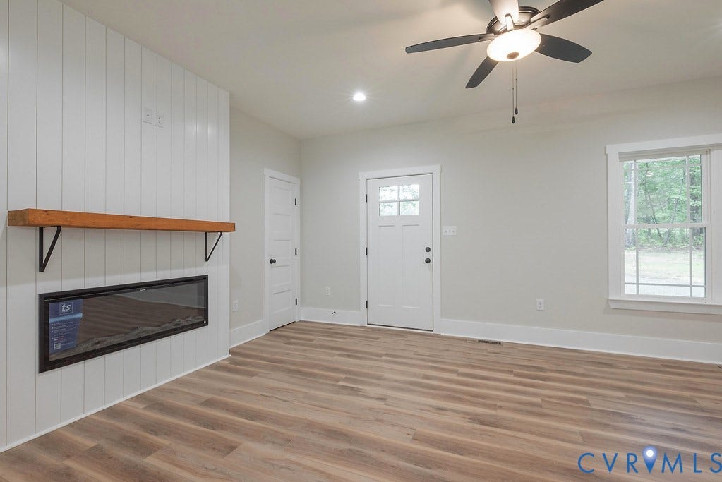 9256 Fredericks Hall Road, Con Creek, Mineral, VA 23117 Spacious interior featuring a contemporary electric fireplace with a wood mantel, wood-finish flooring, a white paneled accent wall, and a ceiling fan with integrated lighting