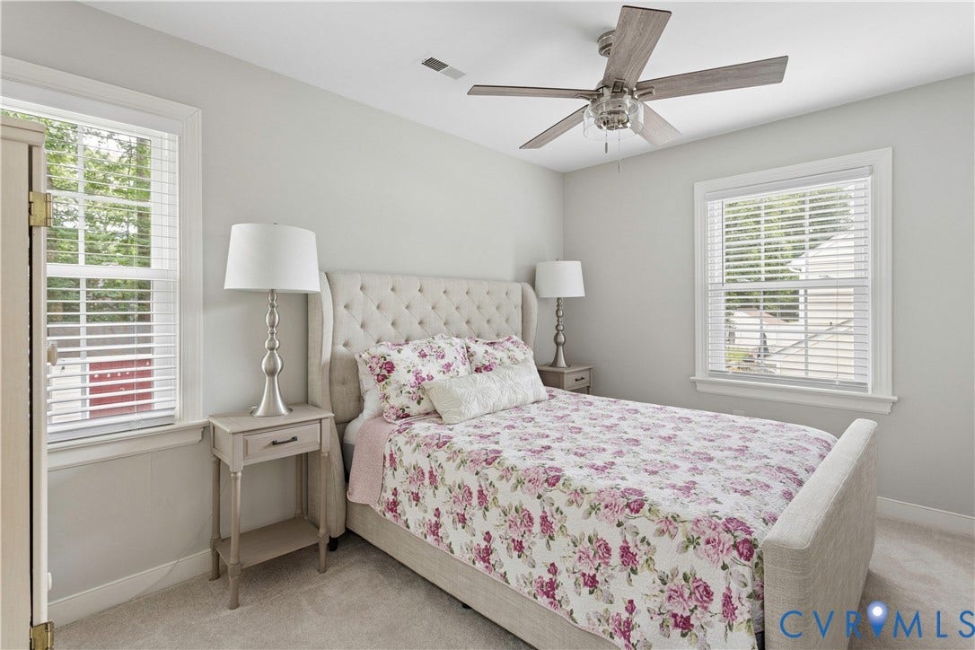 10100 Contessa Court, Glen Allen, VA 23060 Light-filled room featuring a tufted upholstered headboard, a ceiling fan with wood-finish blades, two windows with white blinds, and light-colored wall-to-wall carpeting