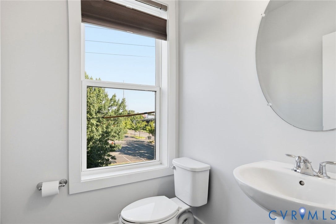 1121 N Lombardy Street, #B, Carver Square, Richmond, VA 23220 Powder Room featuring a single-pane window with a roller blind, a white pedestal sink with a chrome faucet, a round frameless mirror, and a white toilet