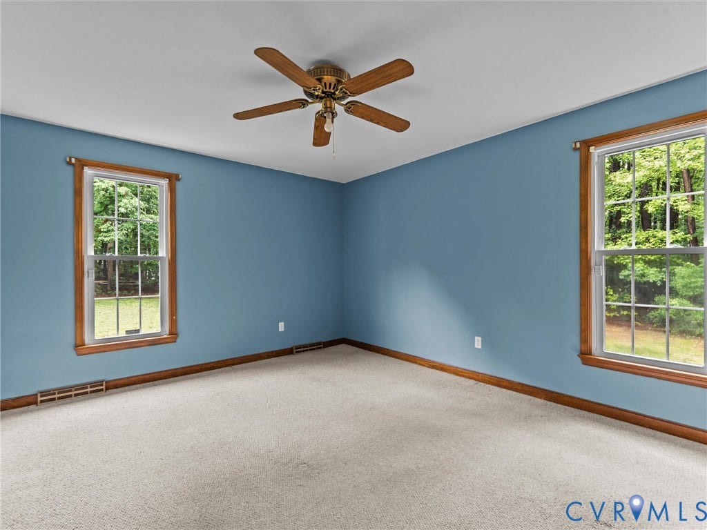 3420 Goodwyn Road, Powhatan, VA 23139 This room features two windows with wood-finish trim, a ceiling fan with light fixture, light-colored carpet, and blue wall finishes.