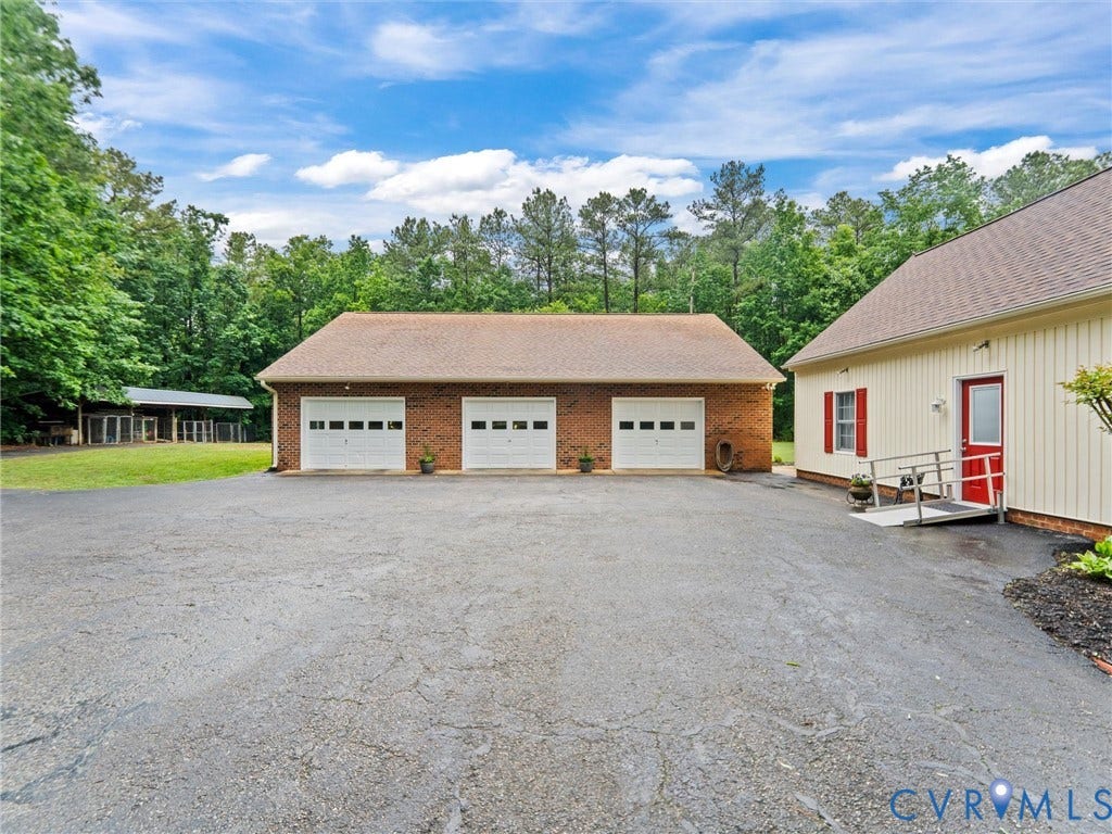 3420 Goodwyn Road, Powhatan, VA 23139 Expansive paved drive with a three-car brick garage, light-colored siding on an auxiliary structure, a red exterior door, and a metal access ramp