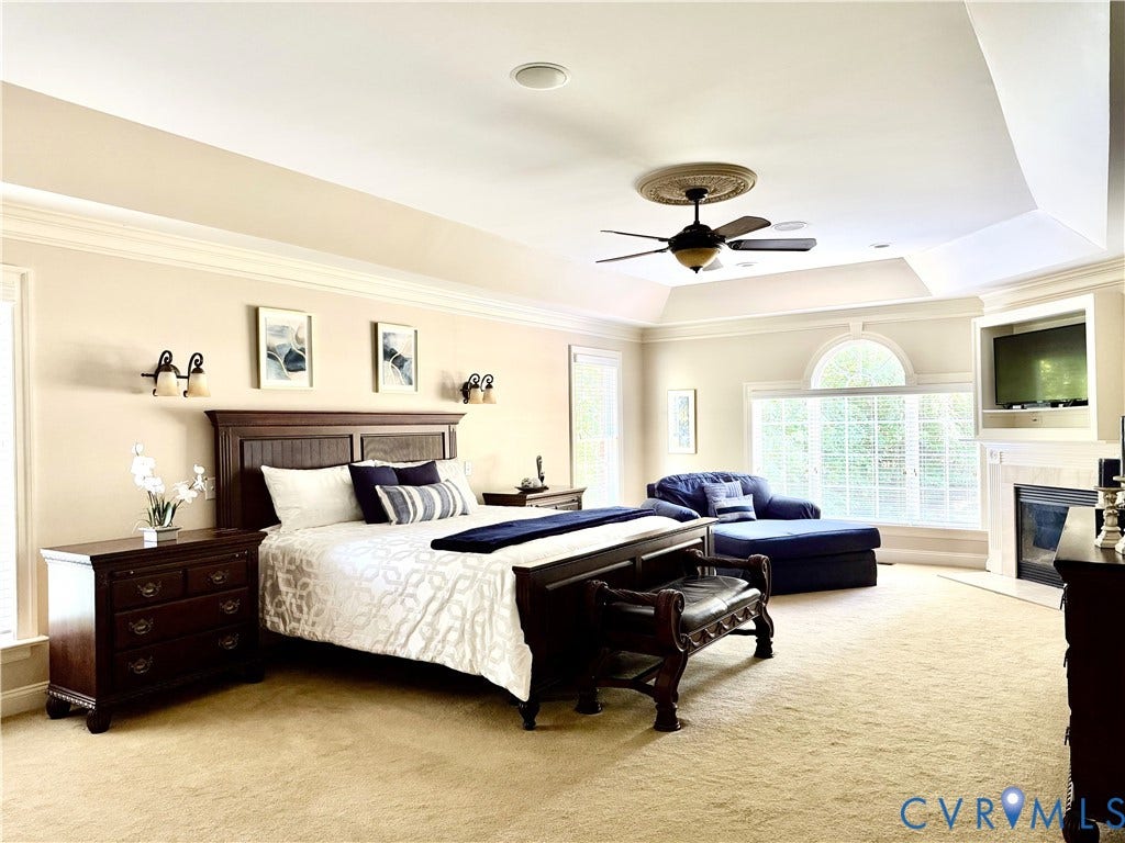 2220 French Hill Terrace, Powhatan, VA 23139 View 1 - Spacious Primary Bedroom with Sitting Area. Notice the Tray Ceiling, Ceiling Fan with Medallion, Gas Fireplace with Television Niche, Crown Molding, Palladian Window, and Ceiling Speakers and Recessed Lights