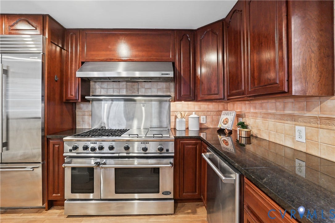 5418 Cary Street Road, Richmond, VA 23226 Kitchen featuring custom cherry-toned cabinetry, stainless steel professional-grade gas range, matching stainless steel hood, built-in refrigerator, and stone countertops with a ceramic tile backsplash