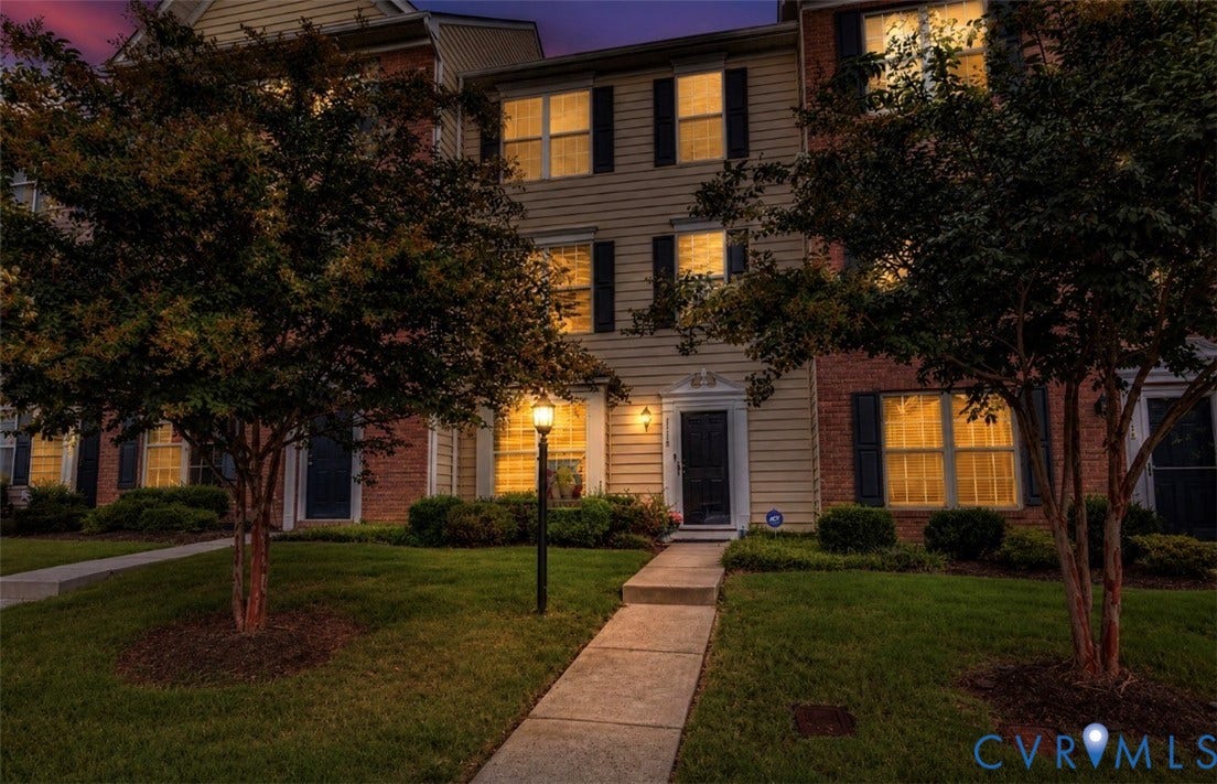 3118 Perdue Springs Lane, Chester, VA 23831 Elegant brick Colonial with illuminated facade featuring multiple tall windows with black shutters, manicured lawn with mature trees flanking a brick walkway, and landscape lighting that highlights the architectural details and grounds