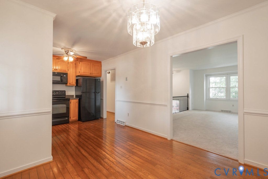 Hardwood flooring extending through the foreground, featuring a kitchen with wood cabinetry and black appliances, and an adjacent carpeted room with multiple windows and a railing
