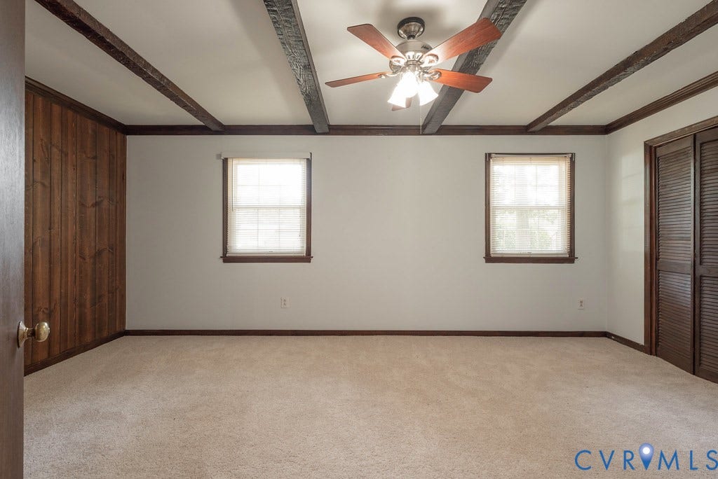 Spacious room featuring exposed ceiling beams, a ceiling fan with light fixture, two windows with wood trim, and a full wall of wood paneling