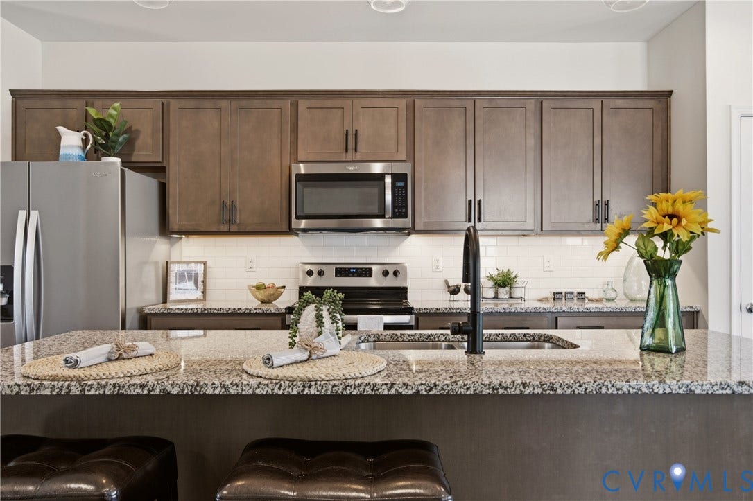 Open-concept kitchen featuring an expansive island with a built-in sink, granite countertops, stainless steel appliances, a subway tile backsplash, and wood-finish cabinetry