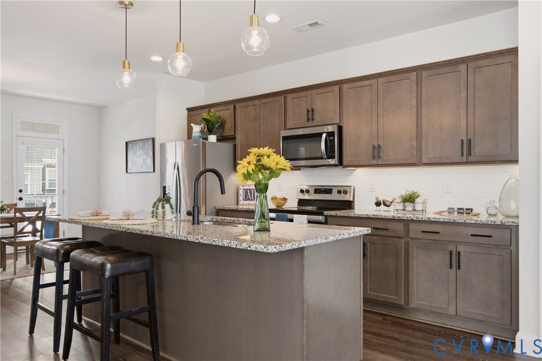 Modern kitchen featuring a spacious island with a built-in sink, granite countertops, stainless steel appliances, contemporary pendant lighting, and wood-finish flooring