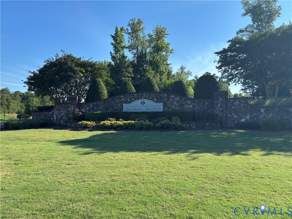 7213 Lyon Taylor Lane, Summer Maple, New Kent, VA 23124 Stone-clad entry monument with a white sign, framed by manicured hedges and mature trees, set against a backdrop of lush green landscaping