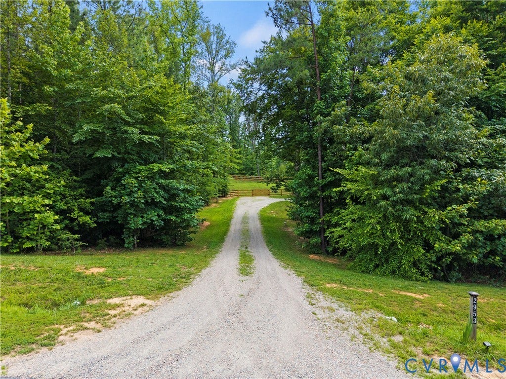 Gravel driveway bordered by lush, mature trees and verdant lawns, leading to a wooden fence