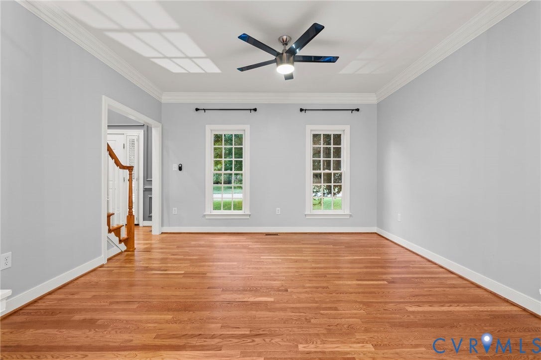 Spacious room featuring wood-finish flooring, two large multi-pane windows, a contemporary ceiling fan, crown molding, and an entryway with a wooden staircase