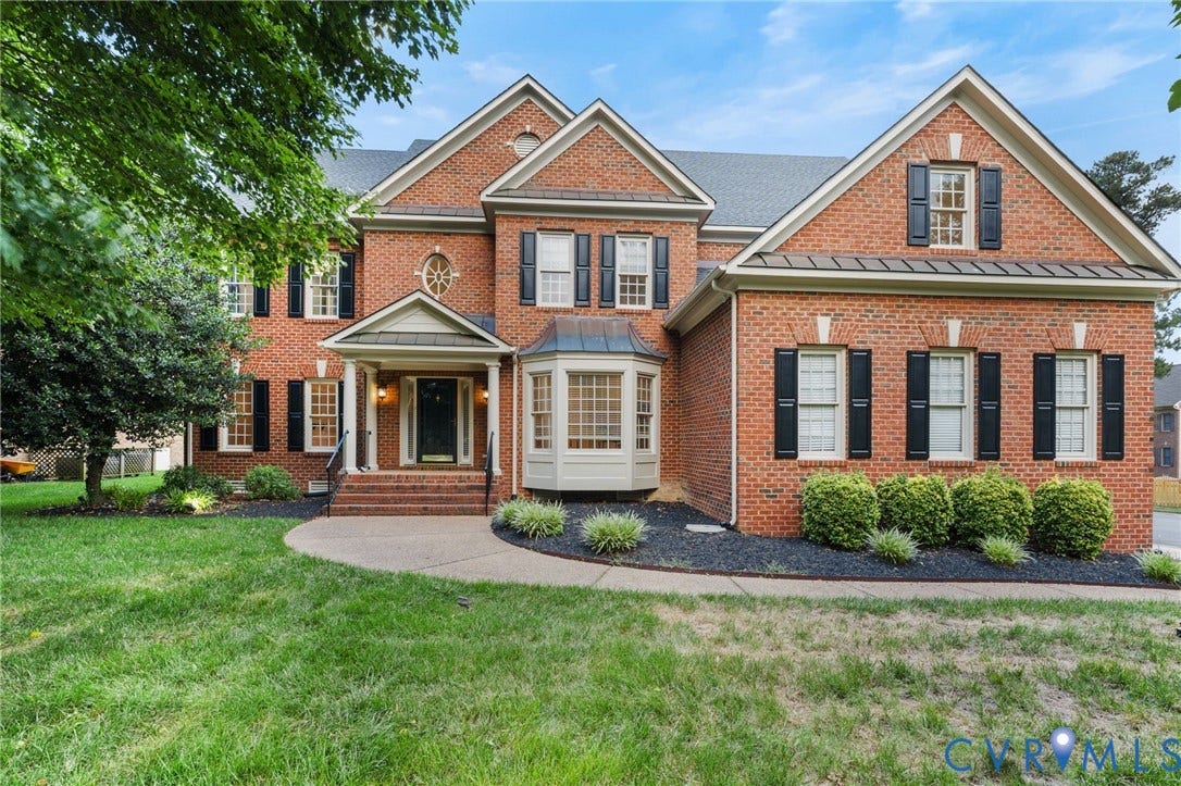 Brick facade residence featuring a covered entry, bay window, black shutters, and mature landscaping