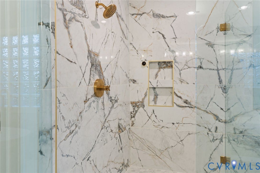 Shower enclosure featuring floor-to-ceiling marbled wall tile with gold-finish fixtures, recessed shelving, and a glass block window